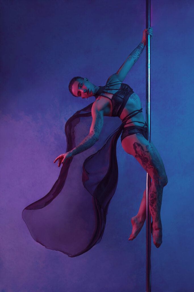 posed pole dancer photo, hanging from a pole, bare foot, black outfit, black flowy cape, blue/purple background