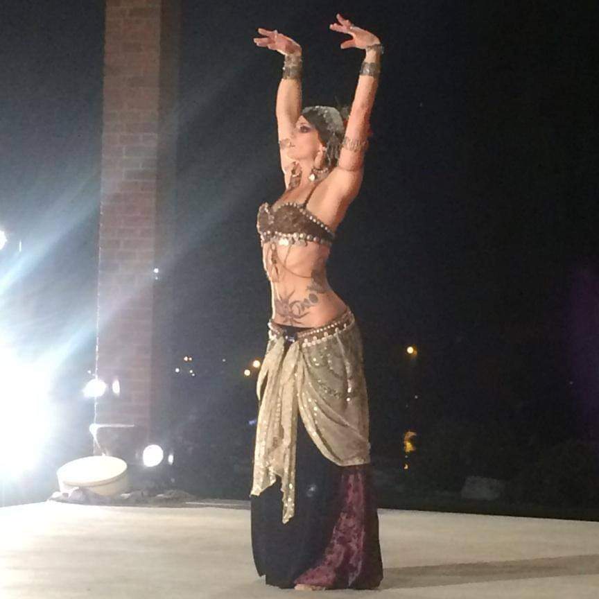belly dancer on stage, arms up in the air wearing a metal headdress and jewelry, a brown bra top, a black skirt, purple harem pants, and cream assuit. bright light shining on her
