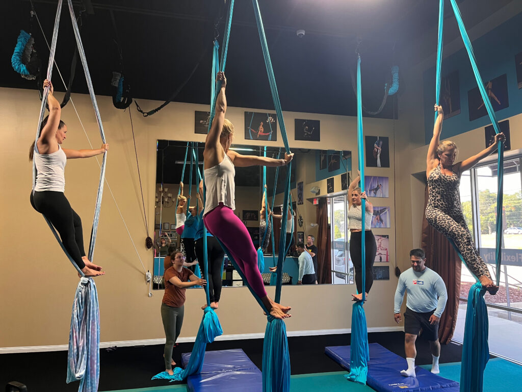 beginner aerial silks class, standing on an egg knot in blue silks, one arm stretched in front of them holding a silk, the other arm grasping the other silk above their head, while leaning their back on that silk