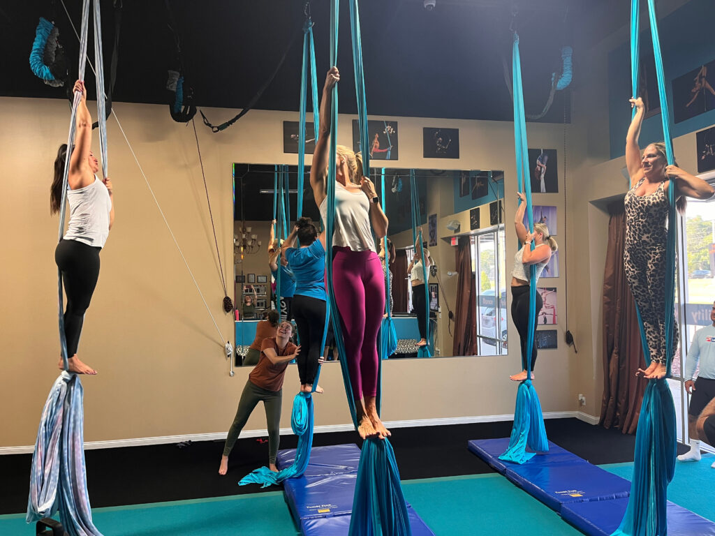 beginner aerial silks class, standing on an egg knot in blue silks, reaching up with one hand